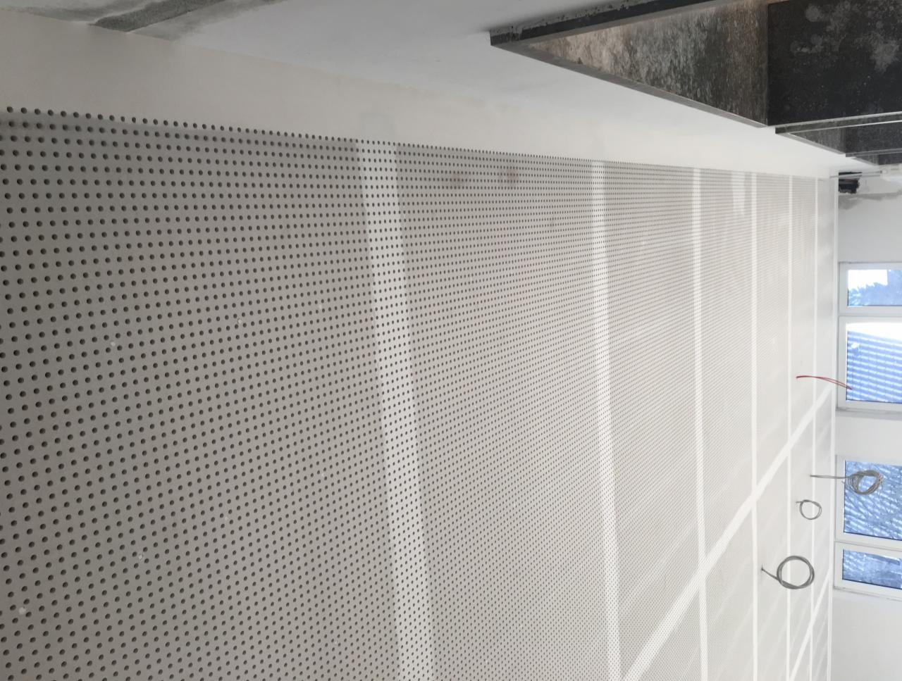 Ściana pokryta perforowaną płytą w trakcie montażu, widoczne okna i fragment sufitu, przewody elektryczne zwisające ze ściany.