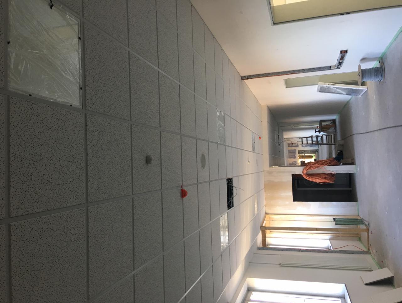 Montaż sufitu podwieszanego kasetonowego w długim korytarzu, widoczne kable i elementy instalacji elektrycznej, częściowo zabezpieczone folią okna.