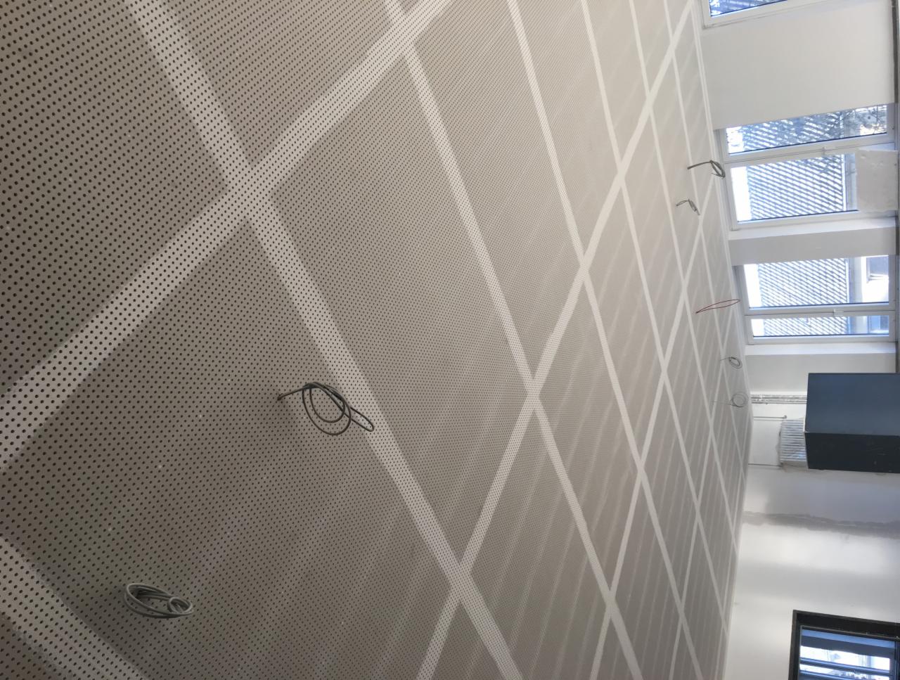 Perforowana, jasnoszara ściana z ukośnymi, białymi pasami, widoczne wystające kable i okna w tle.