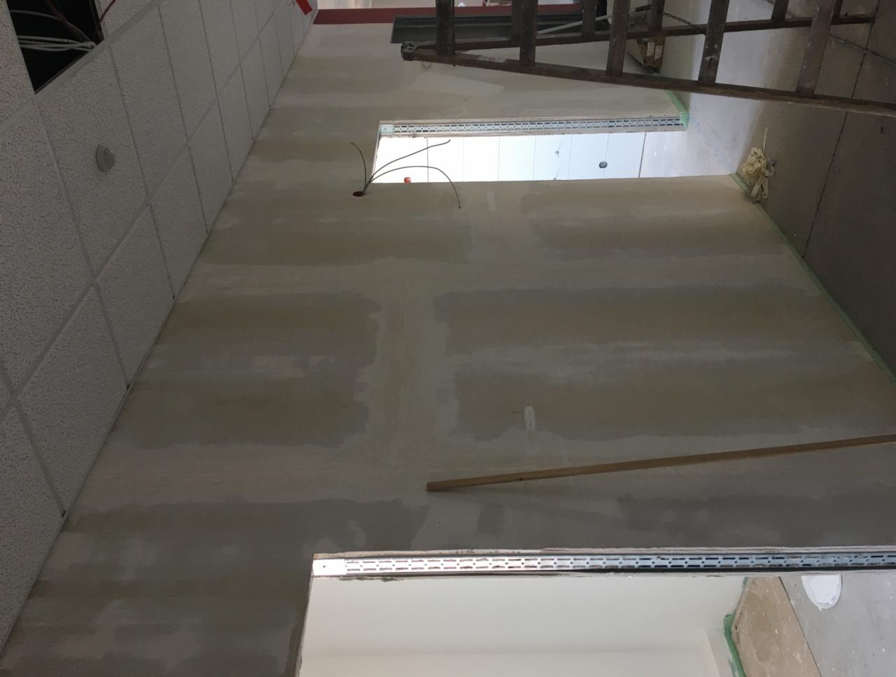 Wnętrze w trakcie prac wykończeniowych: ściany po wstępnym nałożeniu masy, widoczne przewody elektryczne wystające ze ściany, drabina oparta o ścianę, sufit kasetonowy.