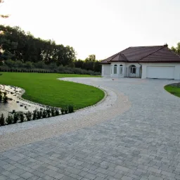 Nowoczesny dom jednorodzinny z zadbanym trawnikiem, podjazdem z kostki brukowej i systemem nawadniania w trakcie instalacji.