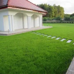 Rozległy, zadbany trawnik przed białym domem z kolumnami i czerwoną dachówką; kamienne płyty tworzą ścieżkę przez trawnik, w tle rząd małych drzewek.