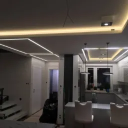 Nowoczesne oświetlenie sufitowe LED w salonie z aneksem kuchennym, widoczne geometryczne kształty i minimalistyczne lampy wiszące.