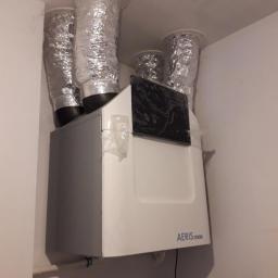 SOLTHERM Włodzimierz Pastuszak - Biało-szara centrala wentylacyjna AERIS z czterema srebrnymi, elastycznymi przewodami wentylacyjnymi podłączonymi do sufitu, widoczne zabezpieczenia folią ochronną.