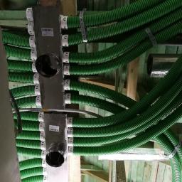 SOLTHERM Włodzimierz Pastuszak - Widok z dołu na system zielonych, karbowanych rur wentylacyjnych podwieszonych pod drewnianym stropem, z metalowymi rozdzielaczami i obejmami mocującymi.