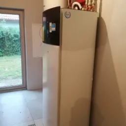 Biała pompa ciepła w rogu pomieszczenia z jasnymi płytkami, widoczne rury i zbiornik na górze, okno z widokiem na zieleń w tle.