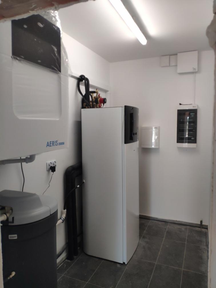 Wnętrze kotłowni z zainstalowaną białą pompą ciepła marki AERIS, szarym zmiękczaczem wody oraz instalacją hydrauliczną z zaworami i rurami w kotłowni wykończonej szarą terakotą.