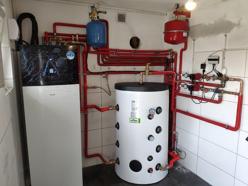 Biała pompa ciepła Daikin z czarnym panelem, zbiornik akumulacyjny Reflex, oraz instalacja z czerwonych rur w pomieszczeniu z białymi płytkami.