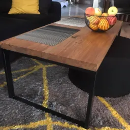 Stolik kawowy z blatem z jasnego drewna na metalowych, czarnych nogach, z owocami w drucianym koszyku i podkładkami na blacie, na tle szarego dywanu z geometrycznym wzorem w kolorze żółtym.