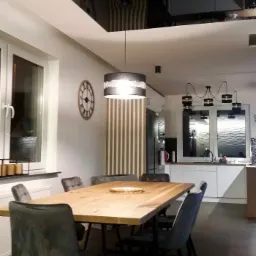 Jadalnia z dużym, drewnianym stołem, krzesłami tapicerowanymi w odcieniach szarości i niebieskiego, oraz widoczną w tle kuchnią z białymi szafkami i oknem z dekoracyjną folią. Na ścianie widoczny...