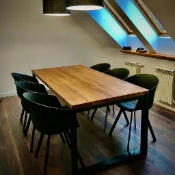 Stół z blatem z jasnego drewna na metalowej podstawie w nowoczesnym wnętrzu poddasza z oknami dachowymi i ciemnymi krzesłami.