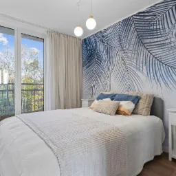 Sypialnia z łóżkiem, balkonem i ścianą ozdobioną fototapetą z motywem liści palmowych w odcieniach błękitu, oświetlona wiszącymi lampami z białymi kulami.