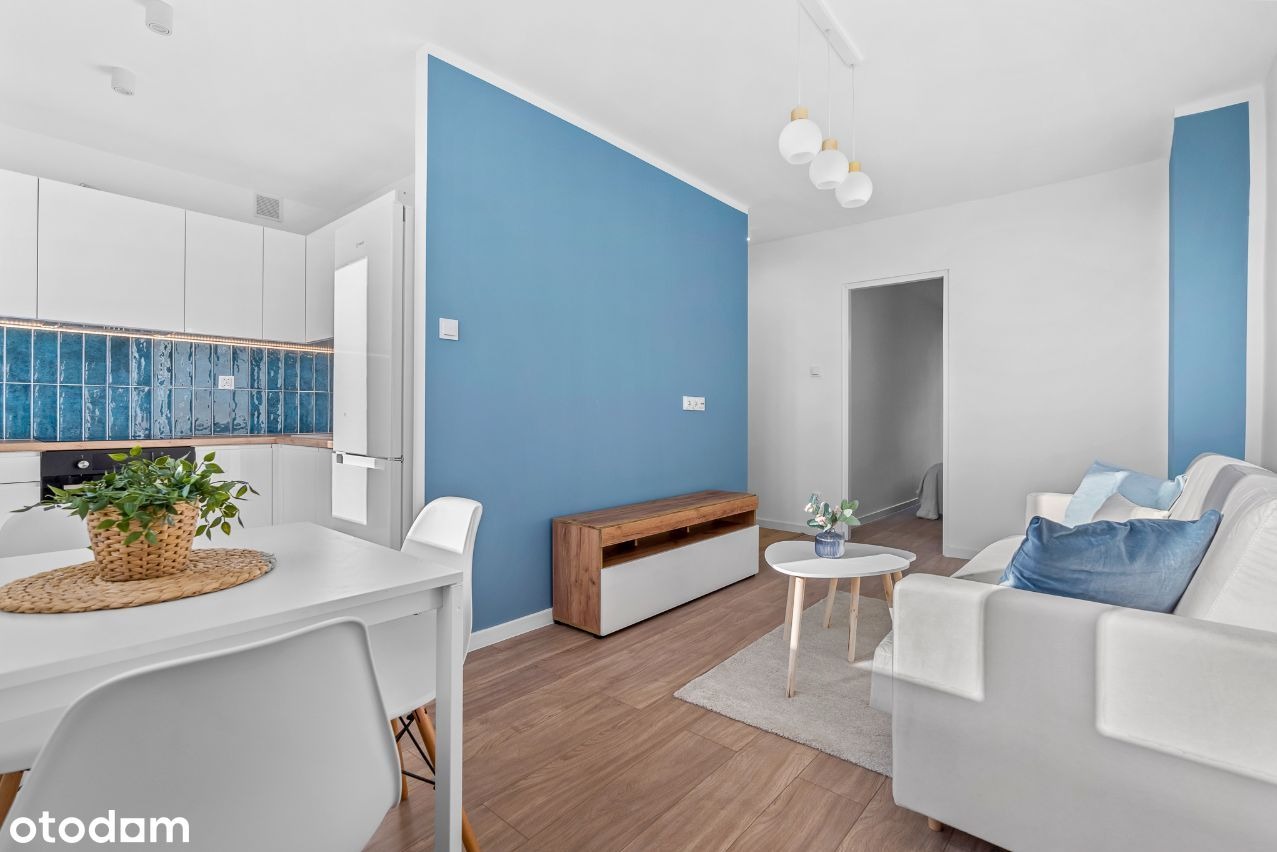 Jasne wnętrze mieszkania po remoncie: białe meble kuchenne z niebieskimi płytkami nad blatem, niebieska ściana w salonie, drewnopodobna podłoga, biała sofa i stół z krzesłami.