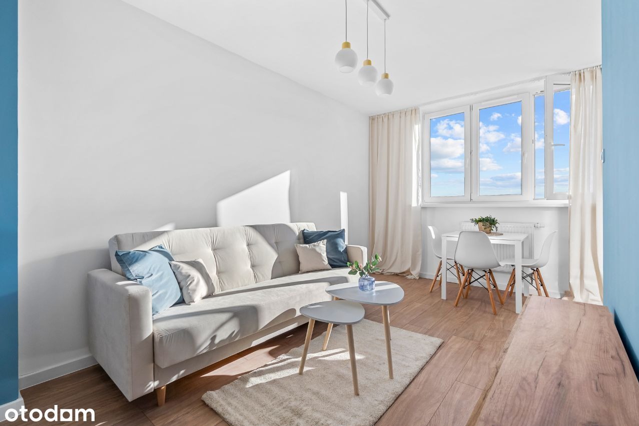 Jasny salon z beżową sofą, drewnianą podłogą i białymi meblami, oświetlony naturalnym światłem wpadającym przez okno z widokiem na błękitne niebo z chmurami.