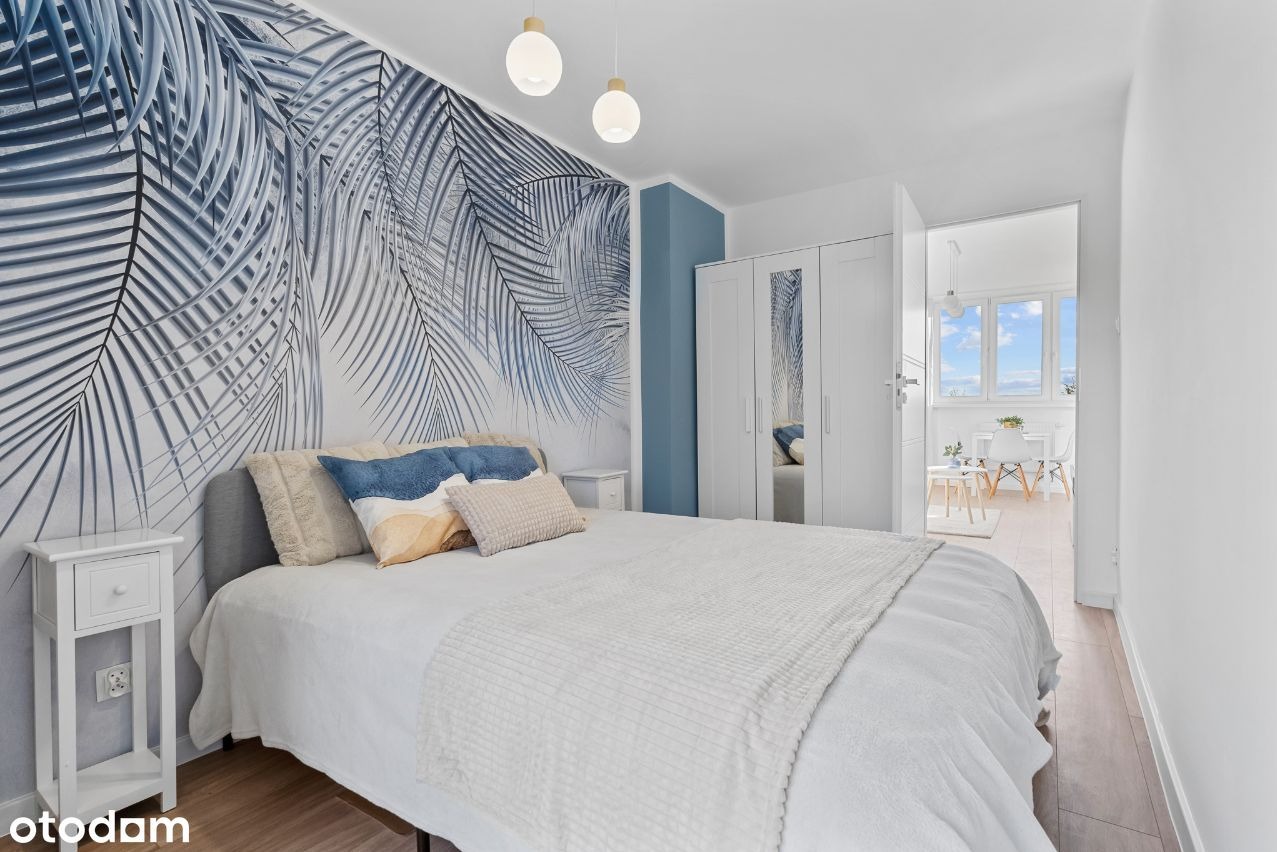 Jasna sypialnia z fototapetą w motywy liści palmowych, białymi meblami i widokiem na balkon z białymi krzesłami.