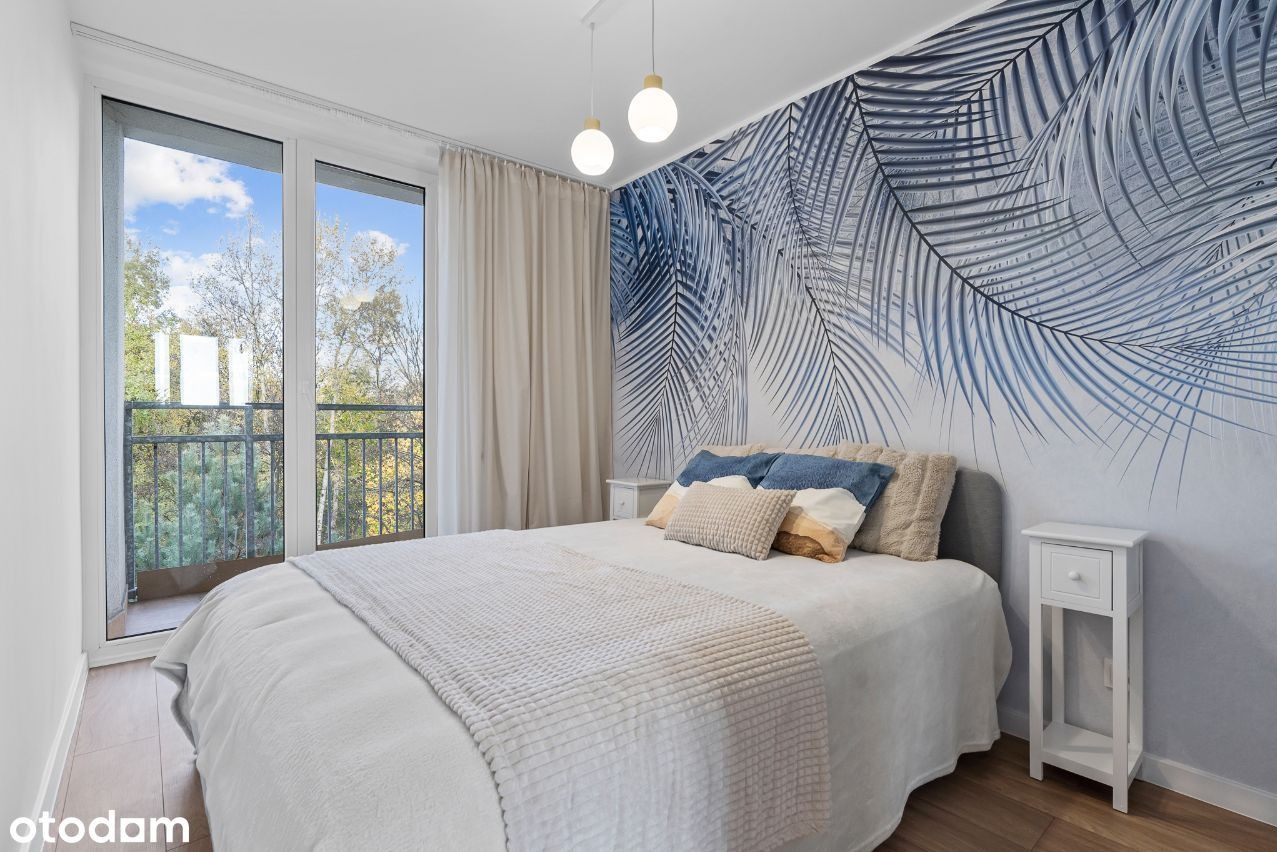 Sypialnia z łóżkiem, balkonem i ścianą ozdobioną fototapetą z motywem liści palmowych w odcieniach błękitu, oświetlona wiszącymi lampami z białymi kulami.