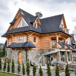 Nowy, ciemny dach z dachówki ceramicznej na drewnianym domu w stylu góralskim z kamienną podmurówką i schodami, widoczni pracownicy podczas prac wykończeniowych.