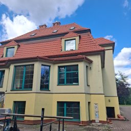 FIRMA DEKARSKO - USŁUGOWA Boro bud - Duży budynek z czerwoną dachówką, licznymi lukarnami i oknami dachowymi. Jasnożółta elewacja z turkusowymi ramami okiennymi. Widoczne rusztowanie na pierwszym planie.