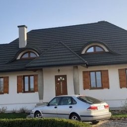FIRMA DEKARSKO - USŁUGOWA Boro bud - Dom z ciemnym, wielospadowym dachem z dachówki, białymi ścianami i brązowymi okiennicami. Dwie lukarny z łukowymi oknami, komin. Przed budynkiem srebrne auto.