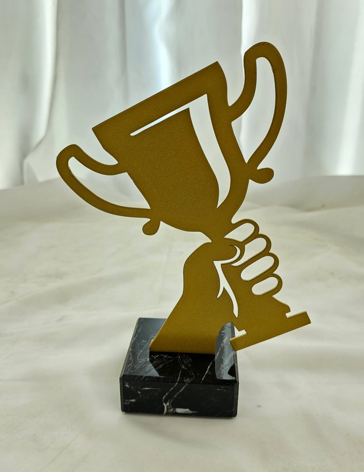 Złoty, metalowy puchar na czarnym, marmurowym postumencie, trzymany w dłoni, na tle białej, delikatnie pofałdowanej tkaniny. Trofeum z bliska, oświetlone miękkim światłem.