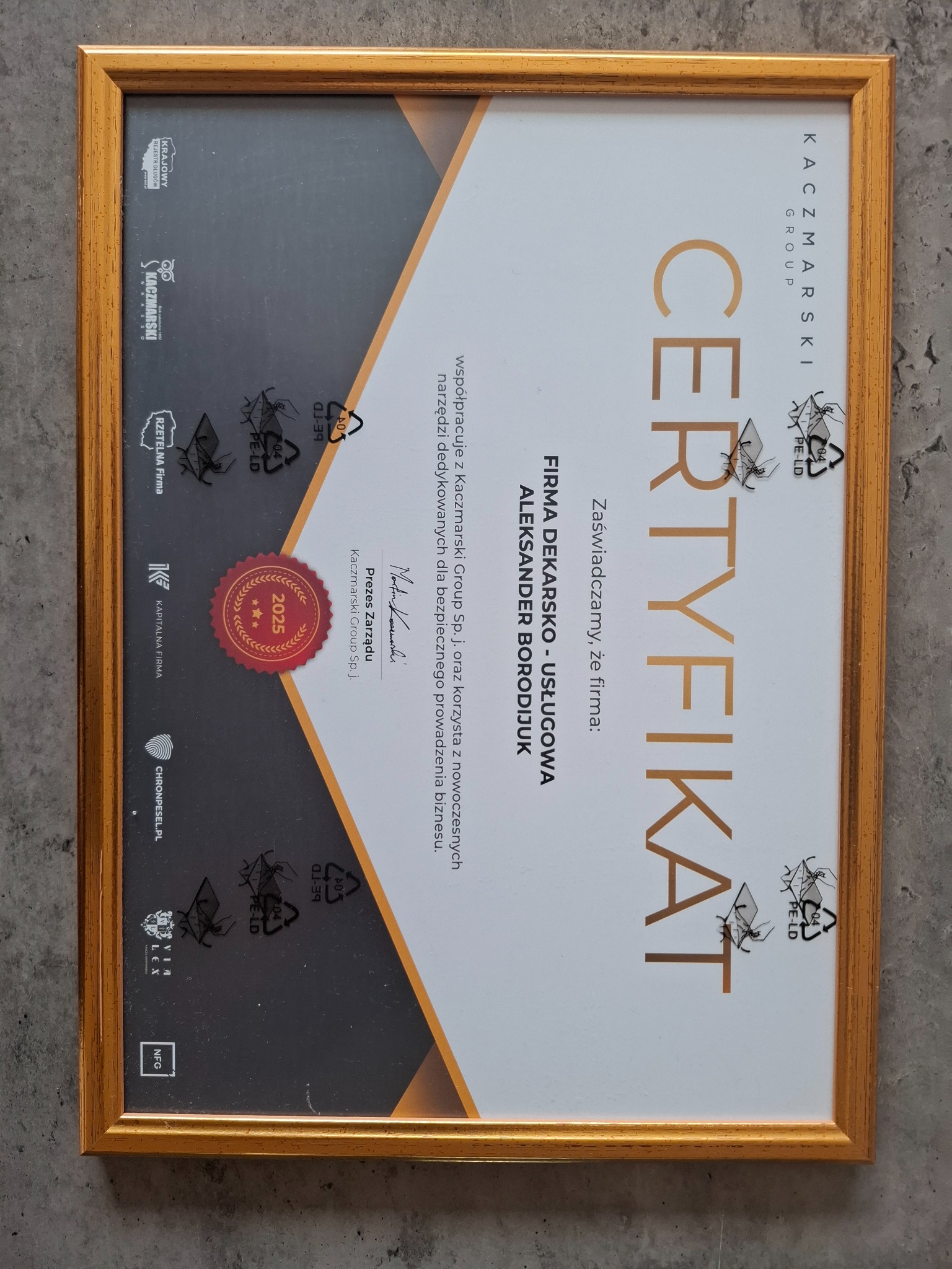 Certyfikat firmy dekarskiej z Suchej w złotej ramie, potwierdzający współpracę z Kaczmarski Group Sp.j. Ważność do 2025 roku. Dokument na szarym tle.