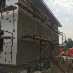 Wznoszony dwukondygnacyjny dom murowany w trakcie ocieplania elewacji, widoczne rusztowania, styropian z fragmentami kleju, szara elewacja, okna bez szyb, pochmurne niebo.