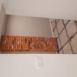 Niski kąt na fragment ściany z ceglaną wstawką i przygotowanym podłożem pod gładzie, widoczny włącznik światła.