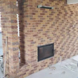 Ściana z cegły klinkierowej w trakcie budowy, z widocznym otworem na kominek i fragmentem otworu drzwiowego, na podłodze widoczne ślady zaprawy, a w tle narzędzia.