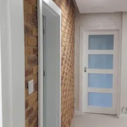 Wnętrze mieszkania z dekoracyjną ścianą z cegły, białe drzwi z szybami i wykończeniami, jasna podłoga.