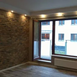 Pokój z dekoracyjną ścianą z cegły, oświetleniem punktowym i drewnopodobną podłogą, widok na balkon przez duże okno.