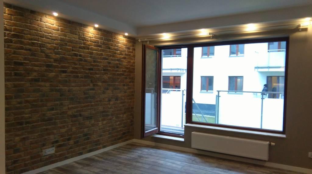 Pokój z dekoracyjną ścianą z cegły, oświetleniem punktowym i drewnopodobną podłogą, widok na balkon przez duże okno.