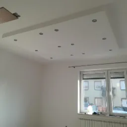 Białe wnętrze z podwieszanym sufitem z wbudowanymi punktowymi lampami, widok na okno z roletami i grzejnik pod nim, fragment niezakończonego sufitu po lewej stronie.