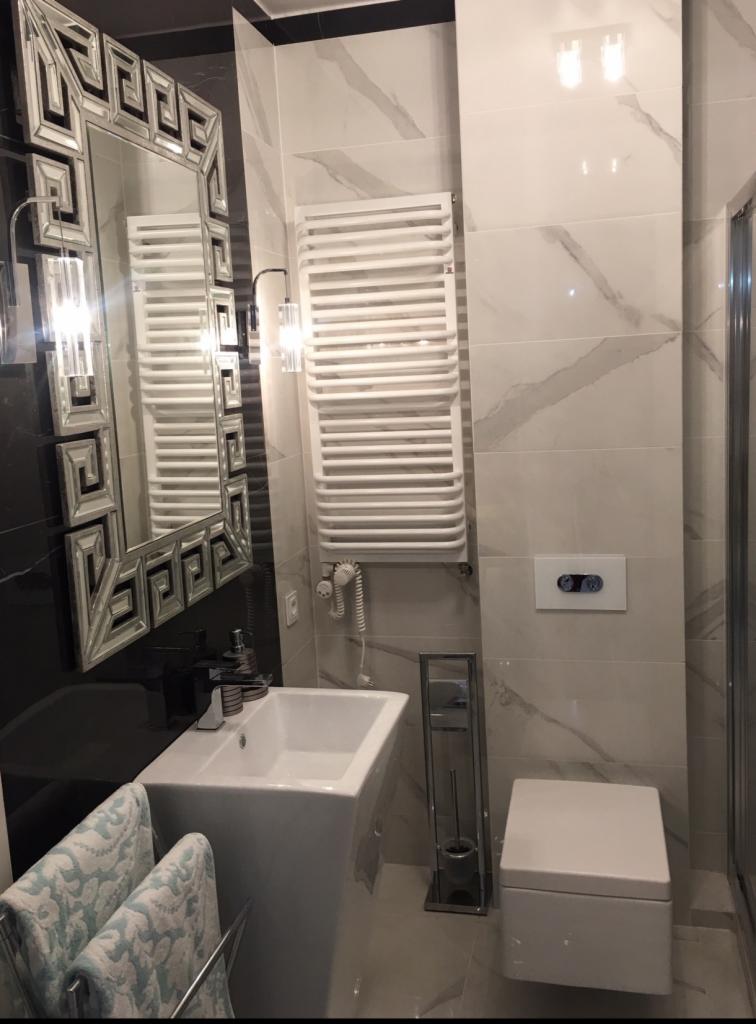 Nowoczesna łazienka z podwieszaną toaletą, grzejnikiem drabinkowym i umywalką na postumencie, wykończona płytkami imitującymi marmur z szarymi żyłami oraz lustrem w dekoracyjnej ramie.