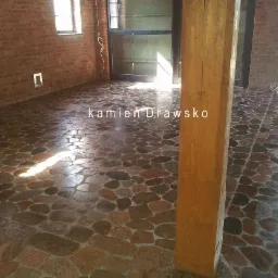 Kamienna podłoga z rzecznych otoczaków w surowym wnętrzu z ceglanymi ścianami, widoczna drewniana kolumna i poziomica.