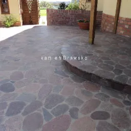 Ułożona z nieregularnych kamieni ścieżka z pojedynczym stopniem, prowadząca do domu z drewnianą bramą i ceglanym murem. Na ścieżce widoczny napis 'kamień Drawsko'.