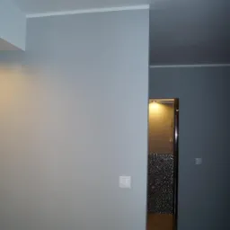 Świeżo pomalowane szare ściany z białym wykończeniem sufitu, widoczny włącznik światła i fragment łazienki za otwartymi drzwiami z dekoracyjną mozaiką.