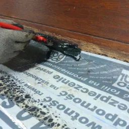 Szczotka z czarną substancją uszczelniającą nakładana na papę izolacyjną przy drewnianej konstrukcji, widoczne ślady wcześniejszego uszczelniania.