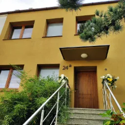 Odnowiona elewacja budynku we Wrocławiu, pomalowana na kolor musztardowy, z drewnianymi oknami i schodami wejściowymi ozdobionymi kwiatami w donicach.