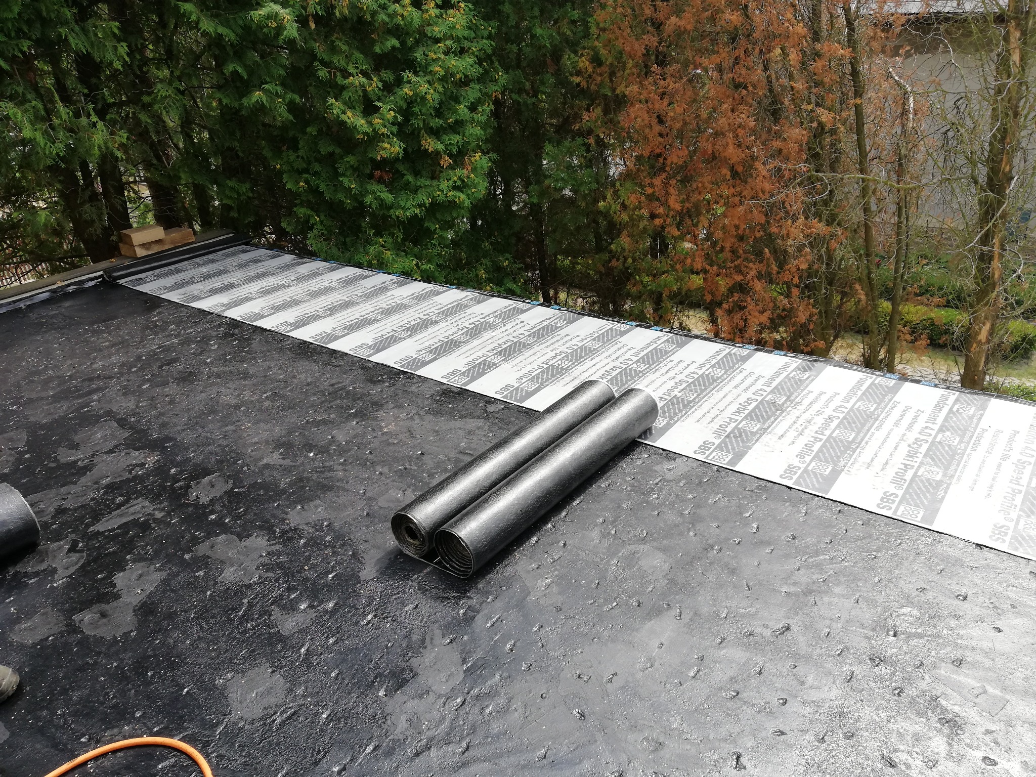 Dwa zwinięte pasy papy leżą na pokrytym bitumem dachu, częściowo zabezpieczonym pasami izolacji z nadrukowanym napisem 'Fundament 4.0 Szybki Profil SBS', w tle widoczne drzewa.