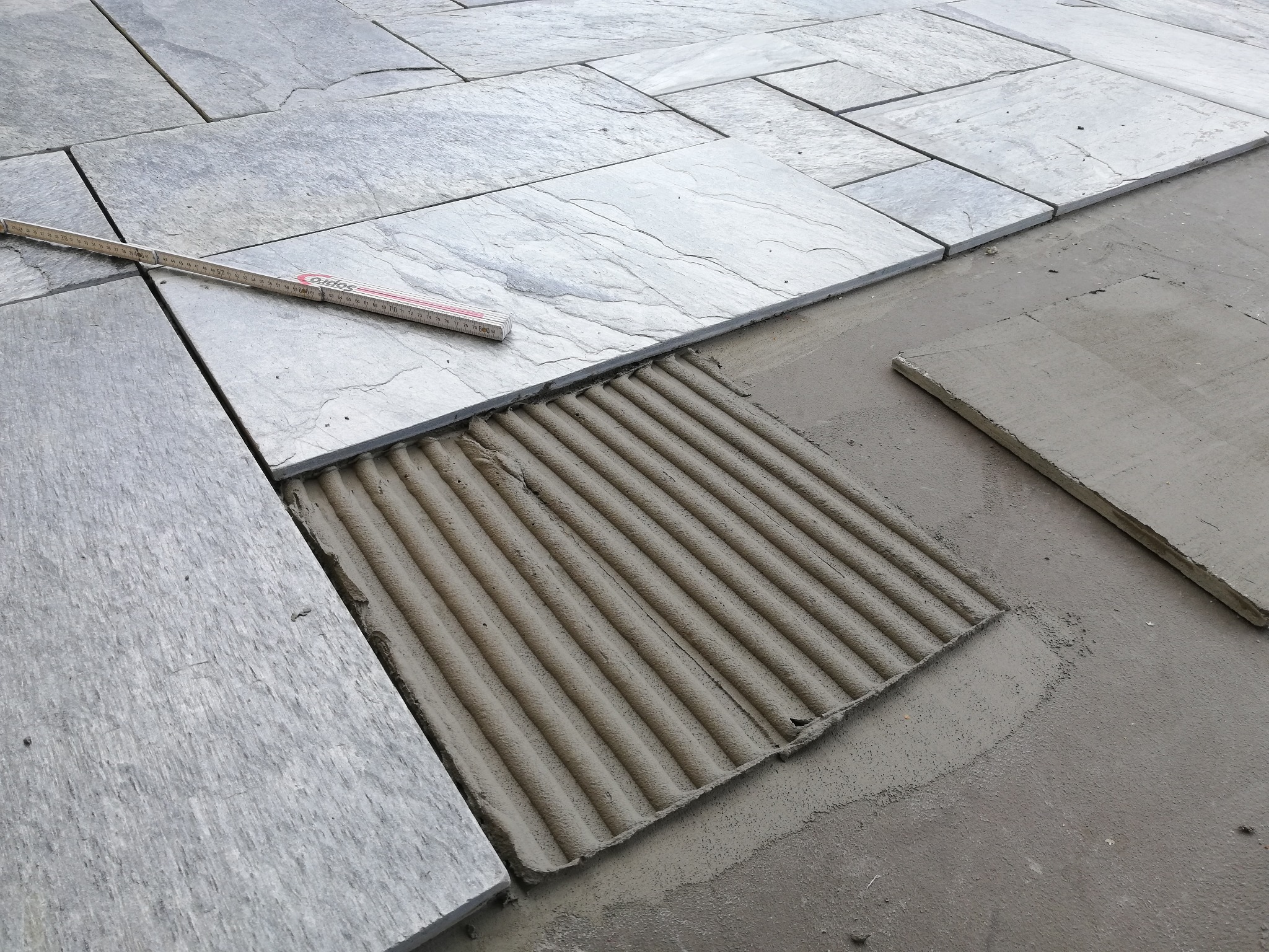 Układanie szarych, prostokątnych płytek na zewnątrz, widoczny klej w paski i miarka stolarska.