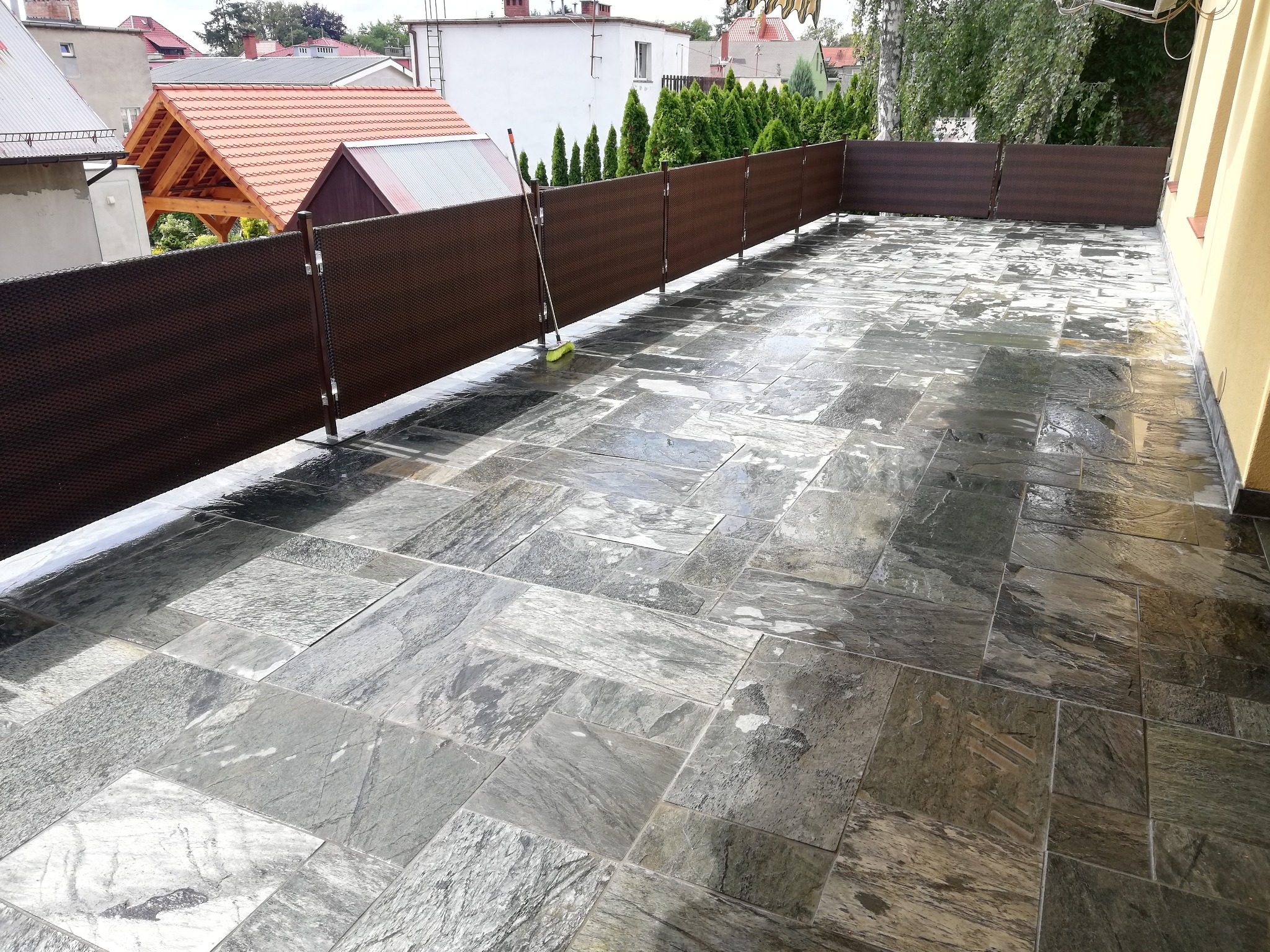 Wyłożony kamiennymi płytami taras z brązową osłoną przeciwwiatrową, na podłodze widoczny ślad po myciu, w tle zabudowa jednorodzinna.