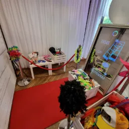 Przygotowana fotobudka z rekwizytami: peruki, kapelusze, okulary i kolorowe boa, odgrodzona czerwonym sznurem od białego tła, z widocznym ekranem dotykowym i napisem 'Paski czy pocztówka?'