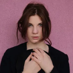 Portret młodej kobiety o niebieskich oczach i brązowych włosach na tle różowej ściany, trzymającej czarny kapelusz w dłoniach.