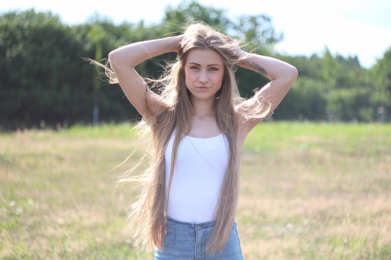 Portret młodej kobiety z długimi włosami na tle zieleni. Modelka w białym topie i jeansowych szortach, z rękami uniesionymi nad głową. Delikatne, naturalne światło.