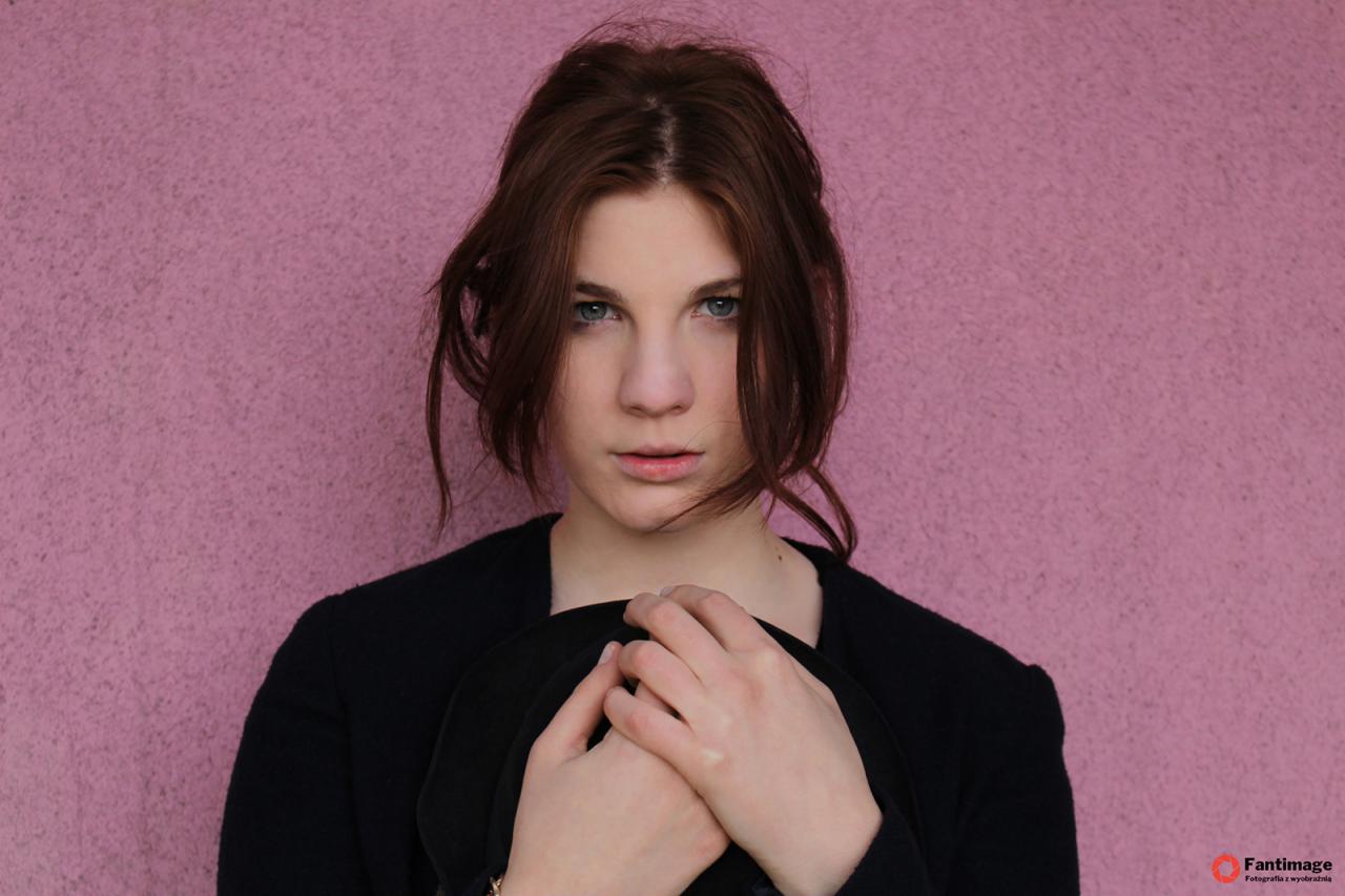 Portret młodej kobiety o niebieskich oczach i brązowych włosach na tle różowej ściany, trzymającej czarny kapelusz w dłoniach.