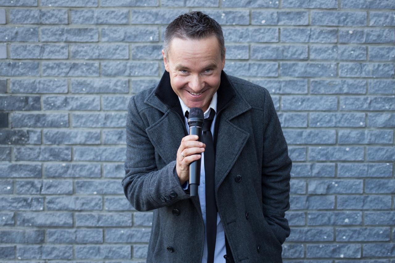 Uśmiechnięty mężczyzna w szarym płaszczu i krawacie, trzymający mikrofon na tle ściany z szarej cegły.