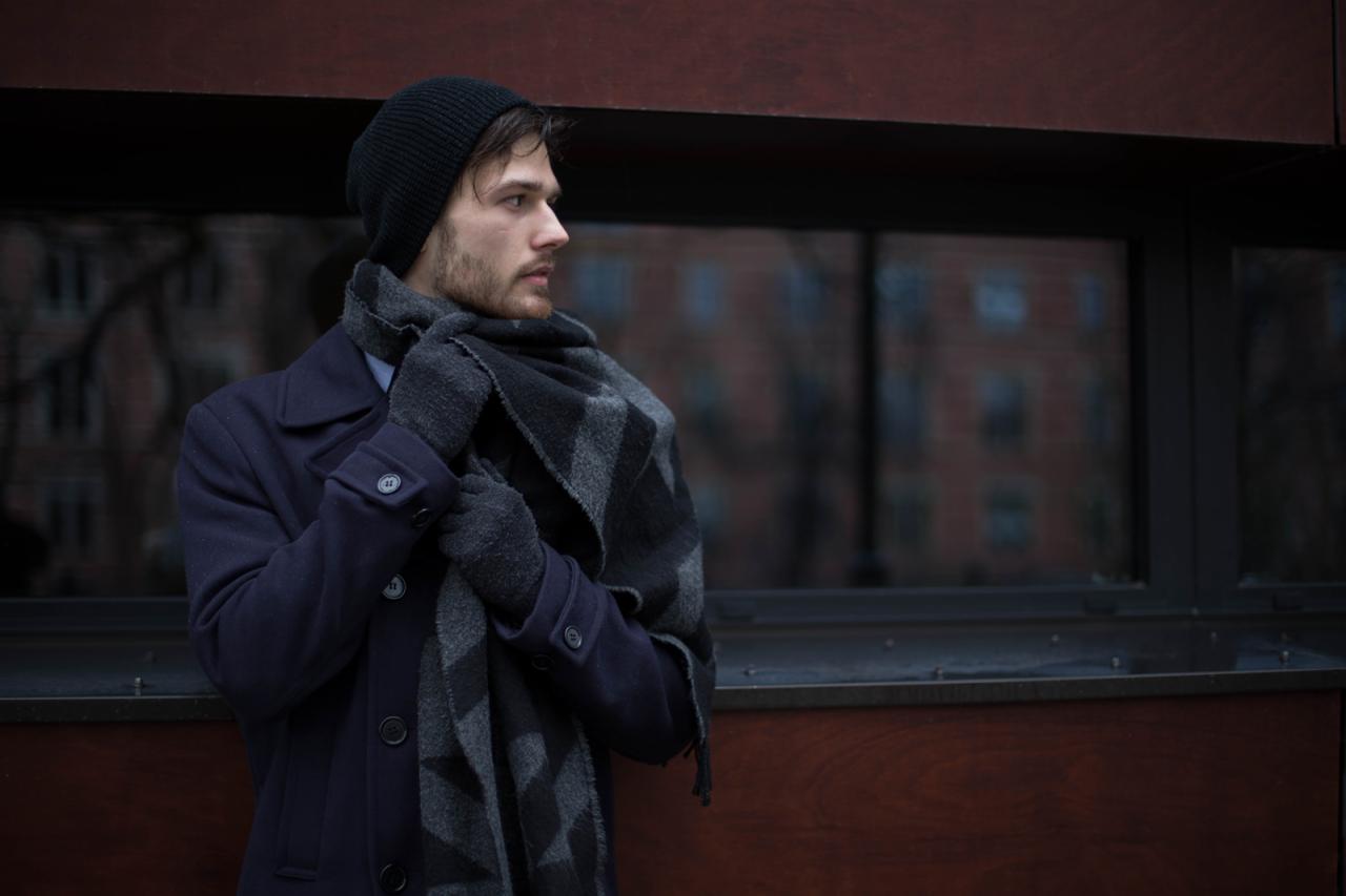 Młody mężczyzna w ciemnym płaszczu, czapce i szaliku w geometryczne wzory, stojący na tle okna z odbiciem budynku, spogląda w lewo.