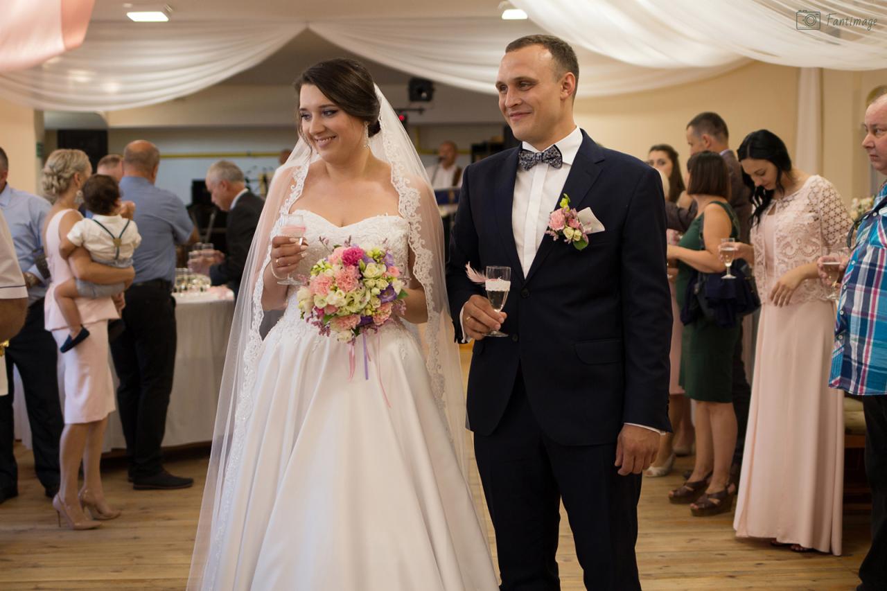 Uśmiechnięta para młoda w tradycyjnych strojach ślubnych, trzymająca kieliszki z szampanem, na tle gości weselnych i dekoracji sali.