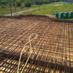 Siatka zbrojeniowa na deskowaniu szalunkowym, przygotowana do zalania betonem, z widocznymi elementami drewnianymi, przewodami i zielonymi bloczkami izolacyjnymi w tle, na tle krajobrazu z drogą...