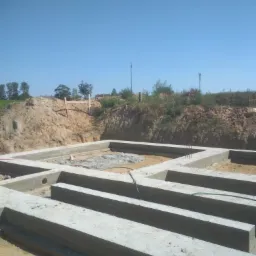 Zalane betonowe fundamenty domu jednorodzinnego z widocznymi otworami i zielonym wężem, na tle wykopu i błękitnego nieba.